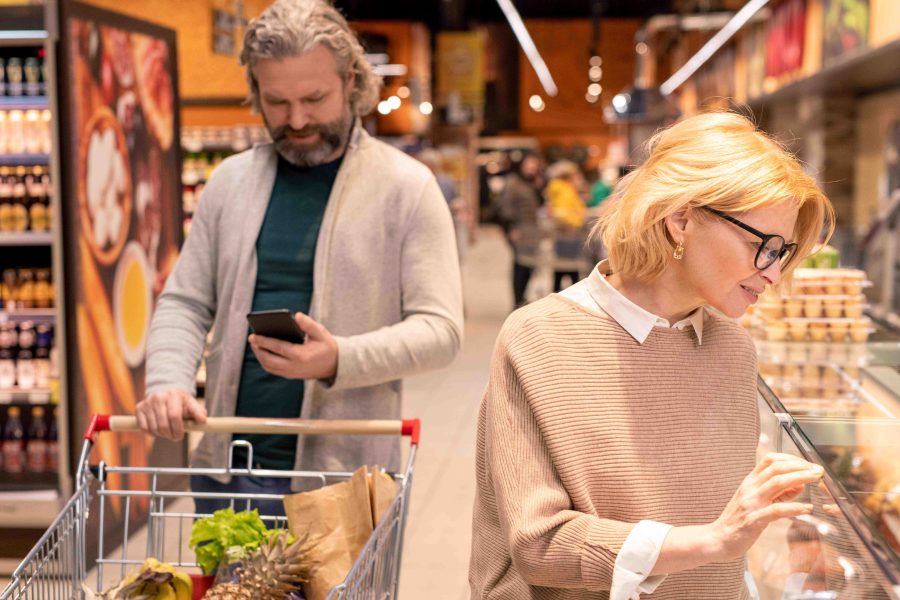 Mature bearded man looking through shopping list smartphone pushing cart while his wife standing by seafood display