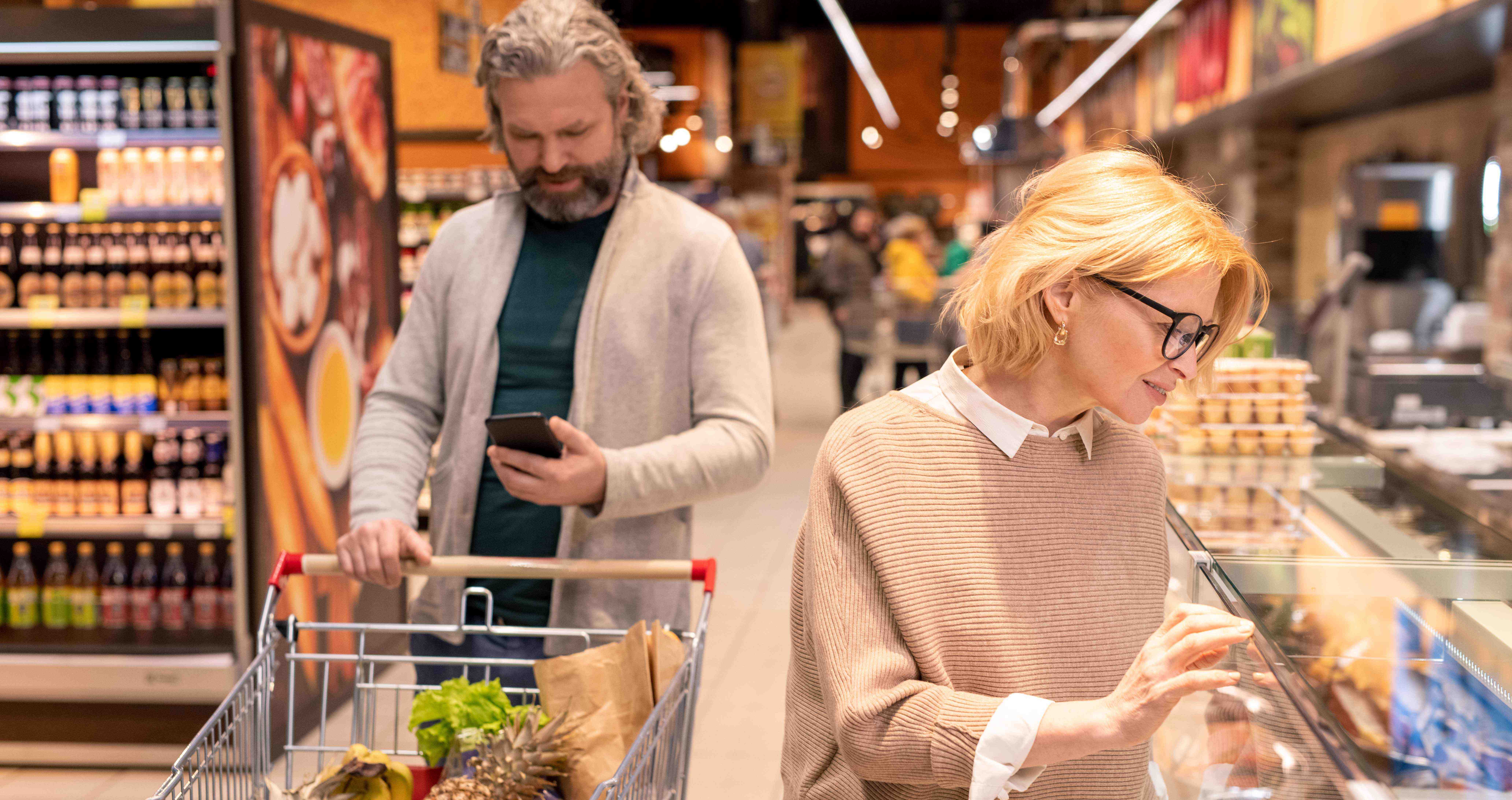 Mature bearded man looking through shopping list smartphone pushing cart while his wife standing by seafood display